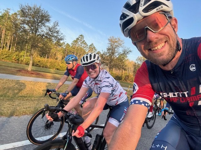 Selfie of three Lodge Ride cyclists, the lead rider in a Shott Performance kit, on a tree-lined road.