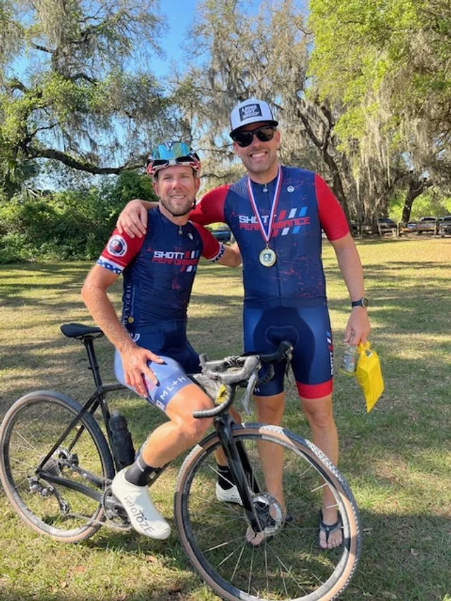 Two SP-kitted athletes posing with their bikes, one wearing a race medal