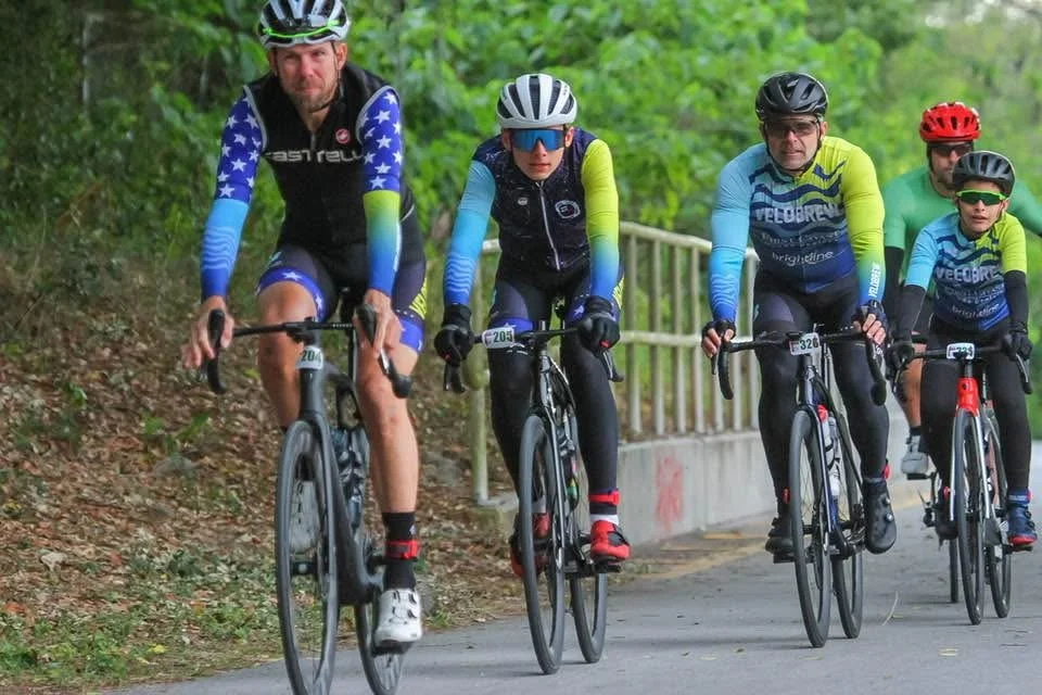 Cyclists wearing numbered race bibs riding together on a shaded path
