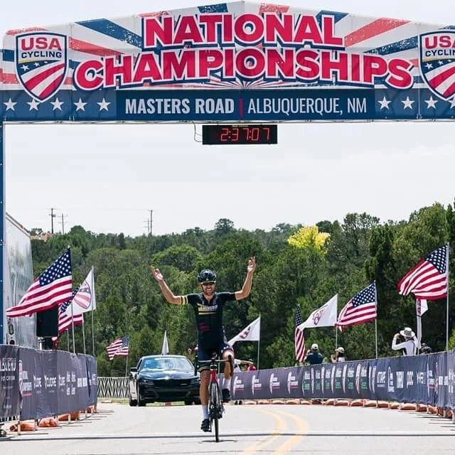 Owen Shott crossing the finish line with arms raised under the USA Cycling National Championships Masters Road arch in Albuquerque, New Mexico
