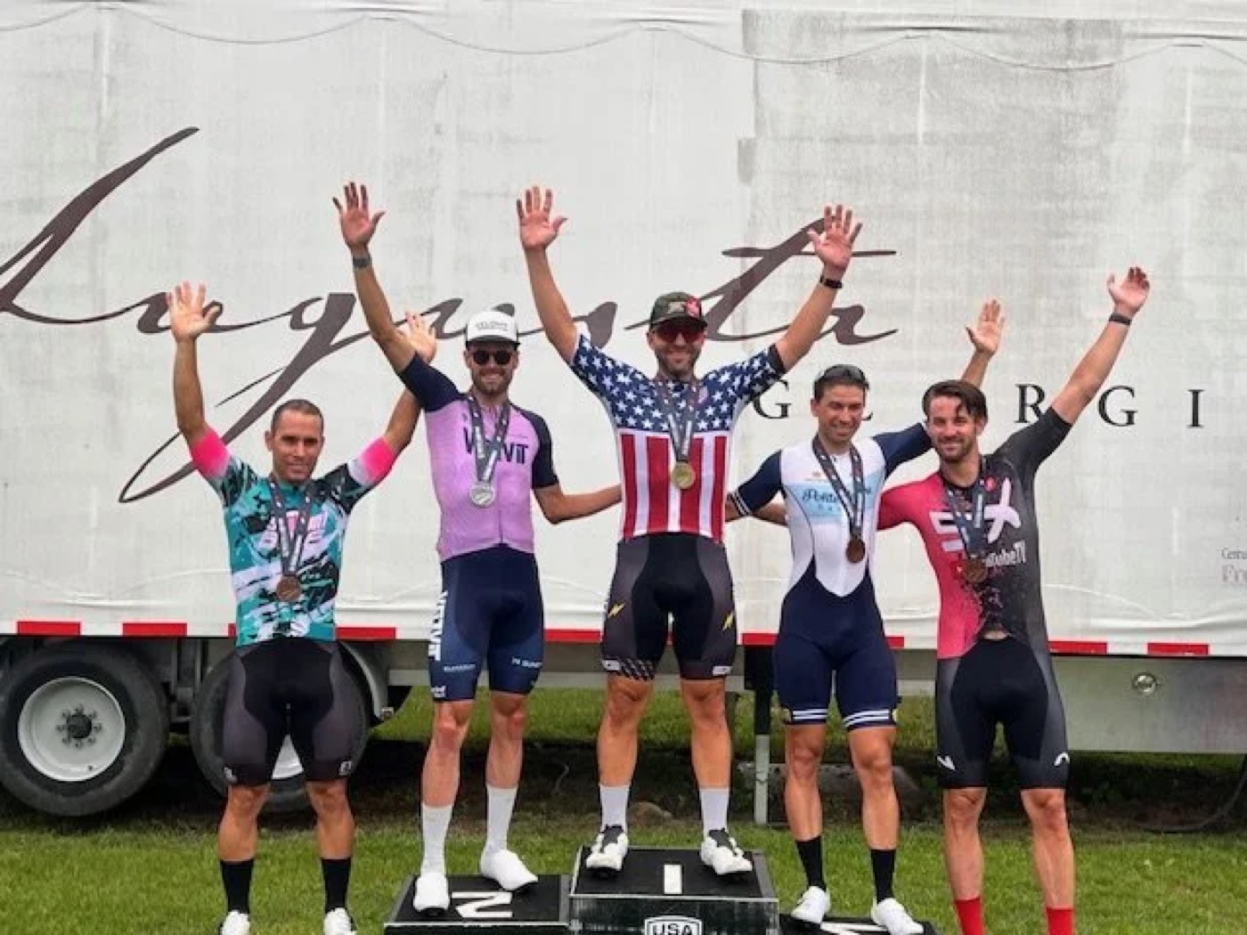 Five cyclists raising their arms together in celebration with medals, in front of an event backdrop, the center rider wearing a stars-and-stripes jersey.