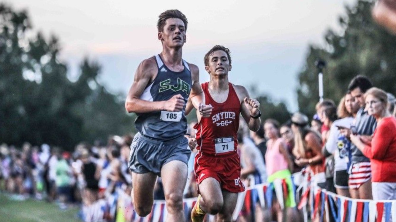 Two high-school cross country runners sprinting side by side toward the finish chute, one in a gray SJA singlet and one in a red kit, spectators and race bunting lining the course in golden-hour light.